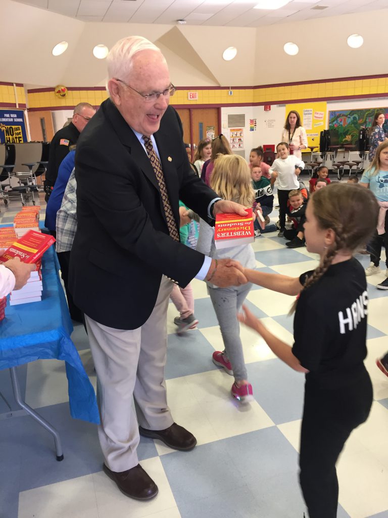 A man in a blue blazer and shirt and tie shakes hands with a third-grade girl with braids while handing her a red dicionary.