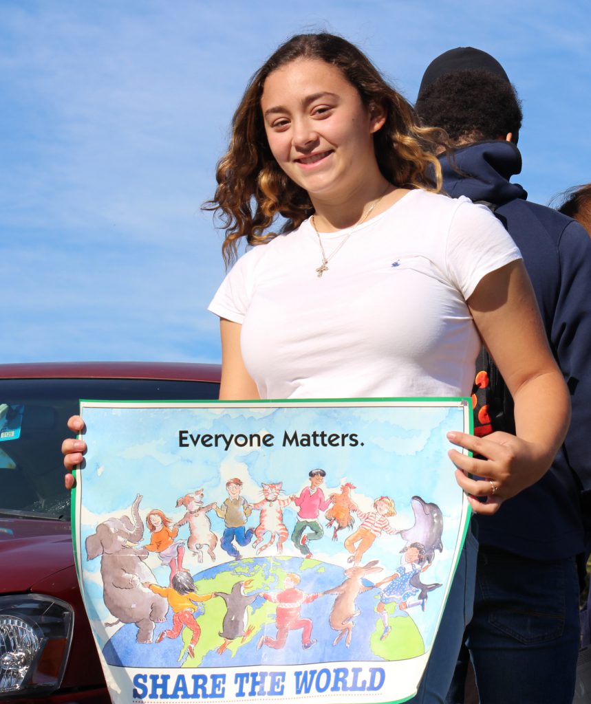 A young woman with dark shoulder length hair wearing a white shirt holds a colorful sign that says Everyone Matters. Share the World.