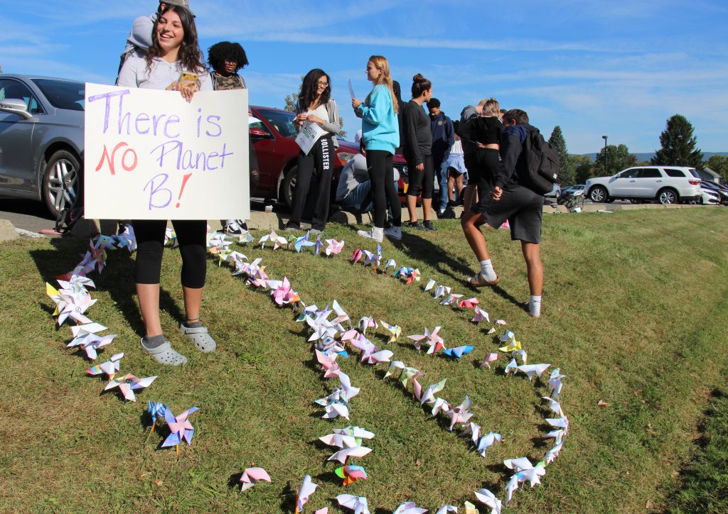 High School students standing on a grassy hill with a peace sign made of small pinwheels. One girl is holding a sign that says "There is no planet B"