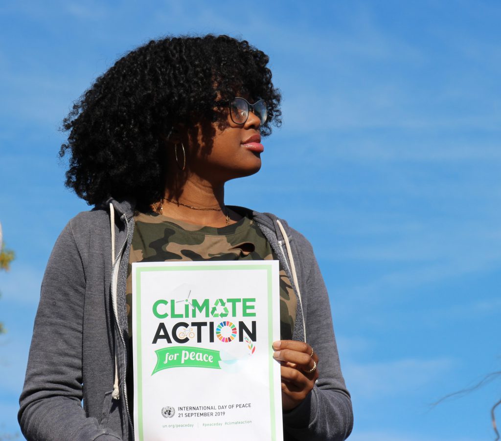 A high school girl with dark hair and glasses wearing a gray sweatshirt holds a poster that says "Climate Action for Peace."