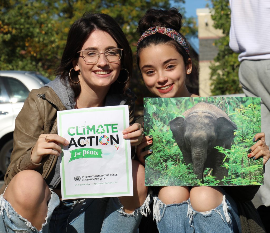 Two high school girls, ,both with long dark hair, hold posters regarding the climate crisis.