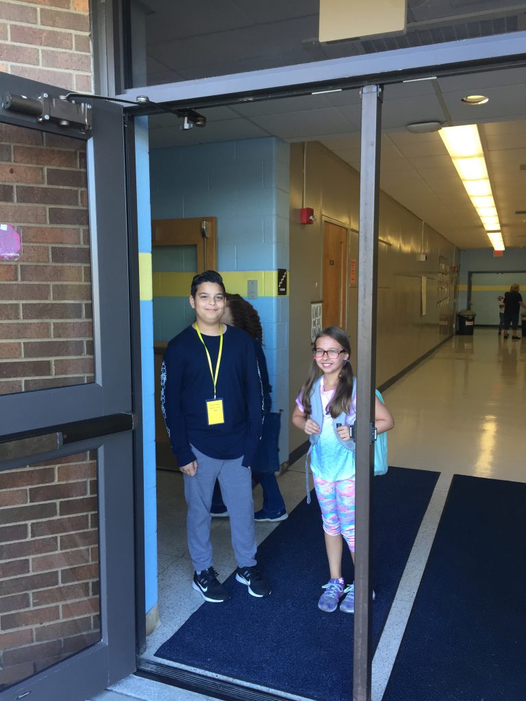 A fifth grade boy stands in a doorway with an elementary school girl. He is a greeter saying hello to everyone who enters