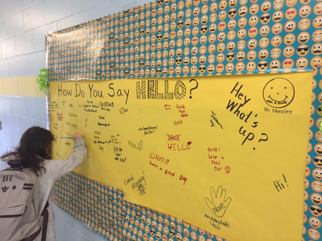 Elementary students sign a large gold poster that is hanging on the wall. They are writing the different ways they say hello