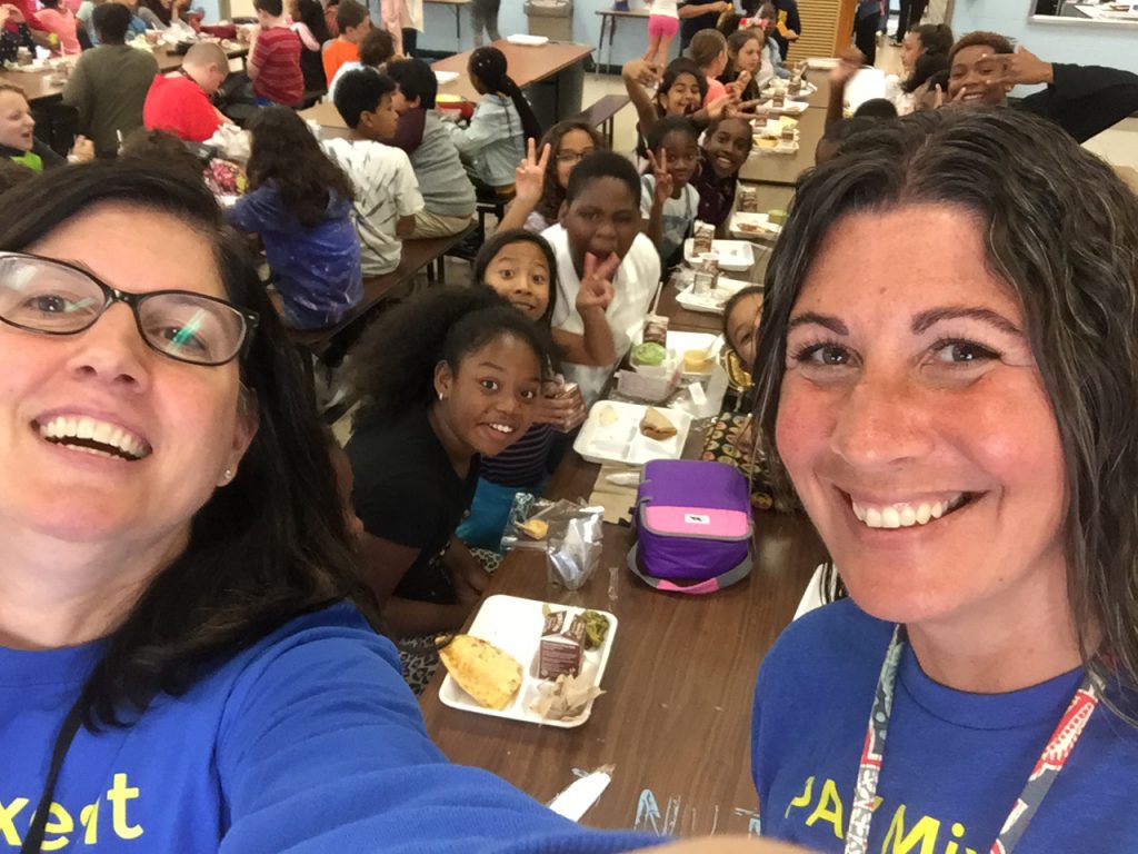 Two teachers stand in front of a long table filled with elementary students eating their lunch