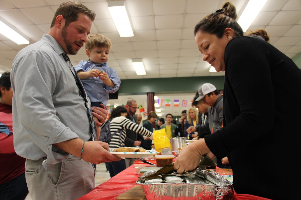 A man holding a toddler holds out his plate as a woman puts food on it. Behind them are lines of people getting food.