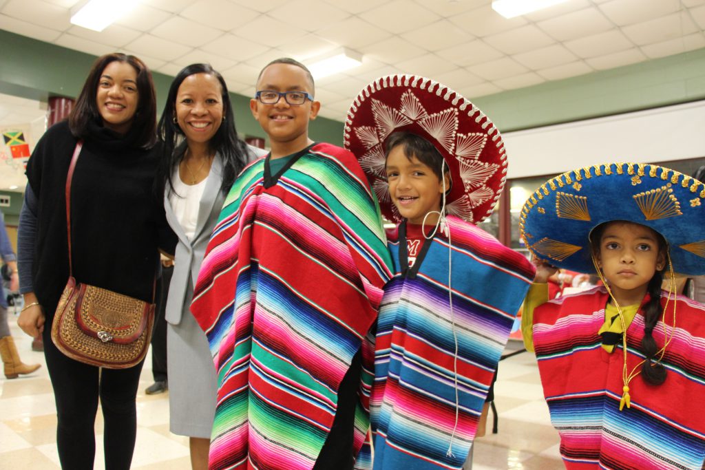 Three children dressed in colorful Mexical garb and large hats stand with two women. All are smiling