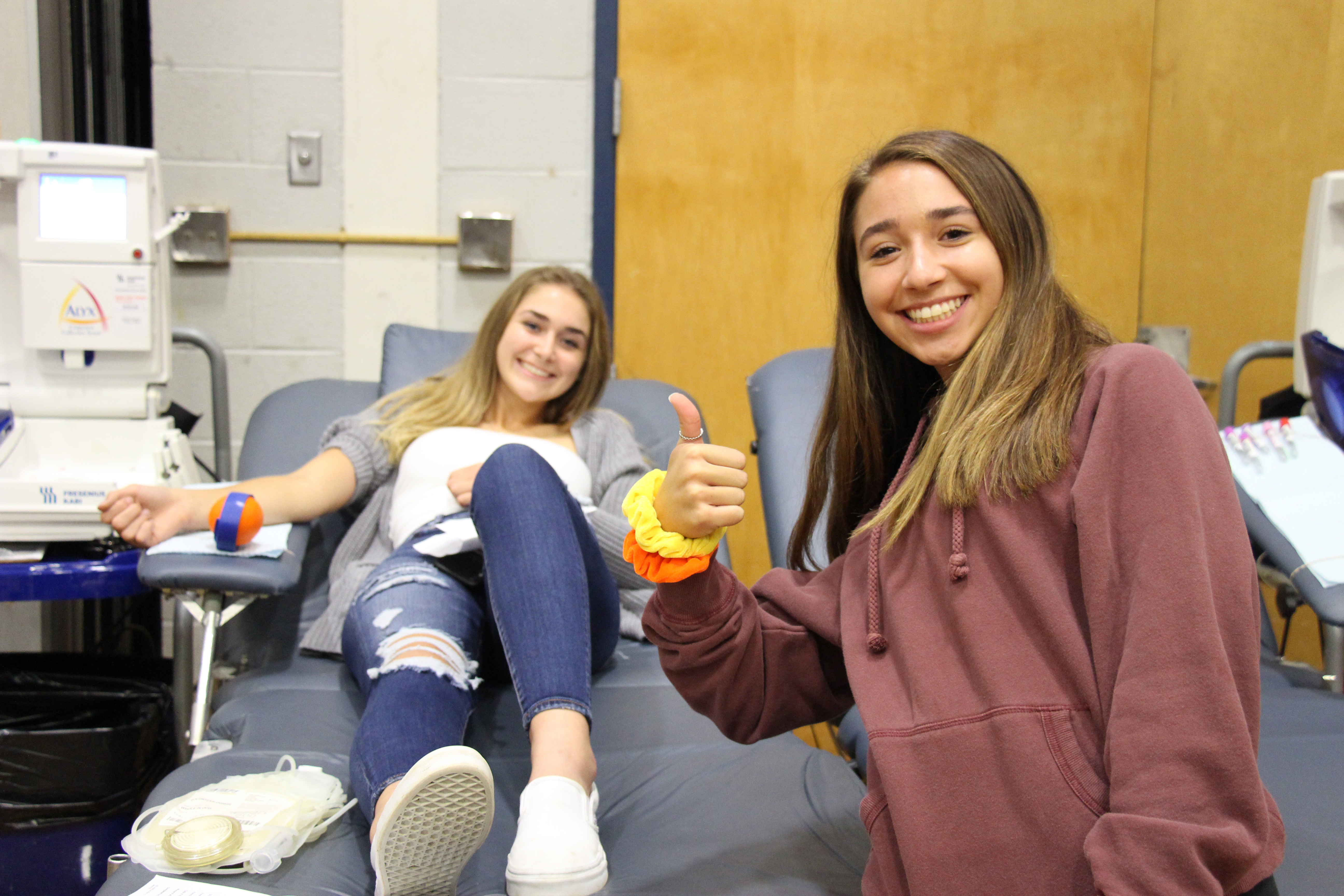 Two young women - one on a gurney giving blood and another in front of her giving a thumbs up.