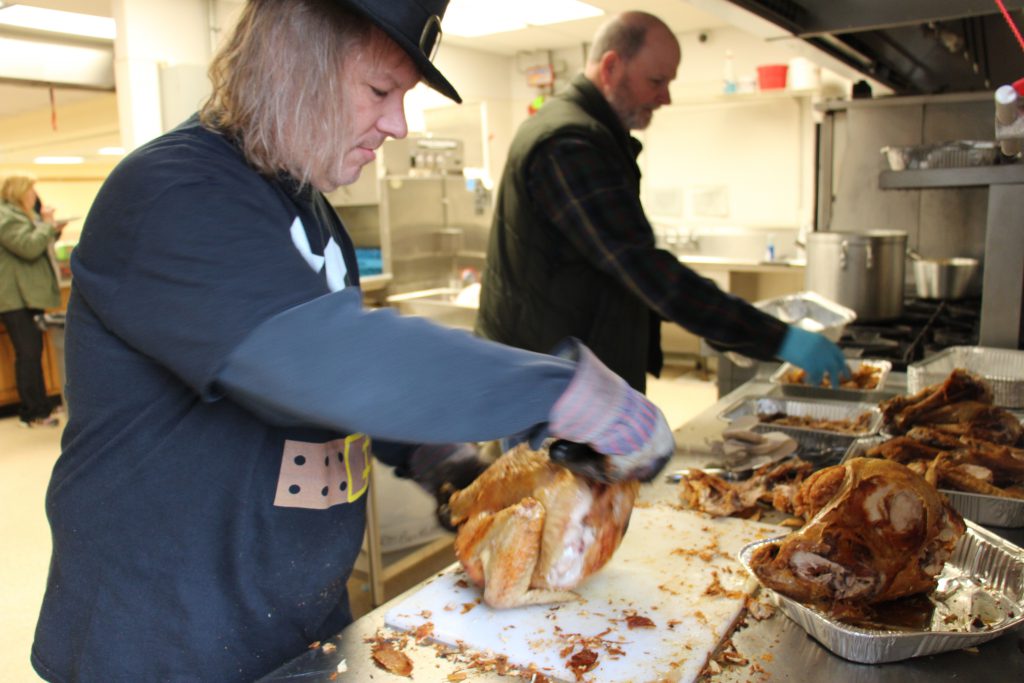 Two men stand at a counter carving tureys. One has long blonde hair and is wearing a pilgrim hat.