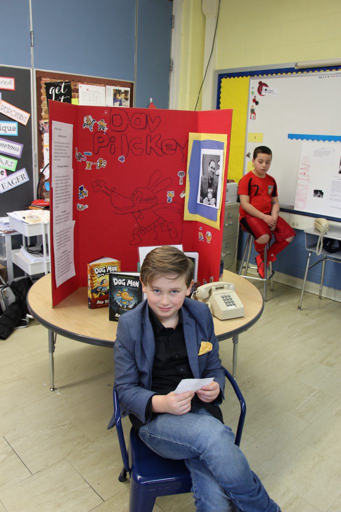 A third-grade boy dressed in a black shirt and blue jacket sits in front of a board with information about his favorite author, Dav Pilkey.