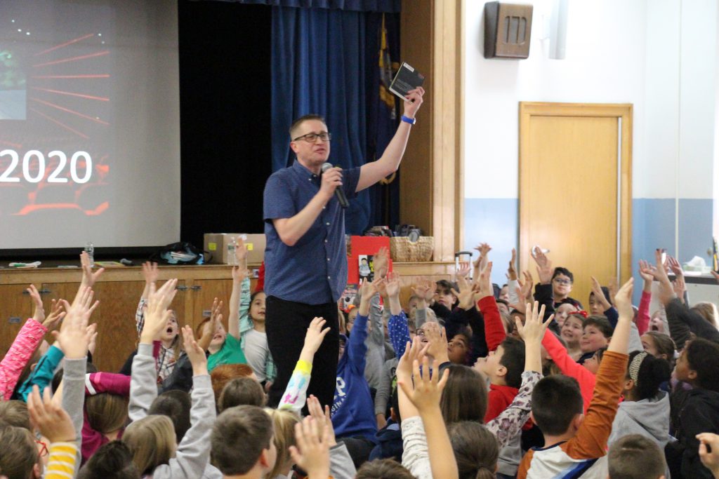 A man in a short-sleeved blue shirt and glasses holds a microphone in one hand and a book in the other, high over his head. Many children sitting on the floor are raising their hands.