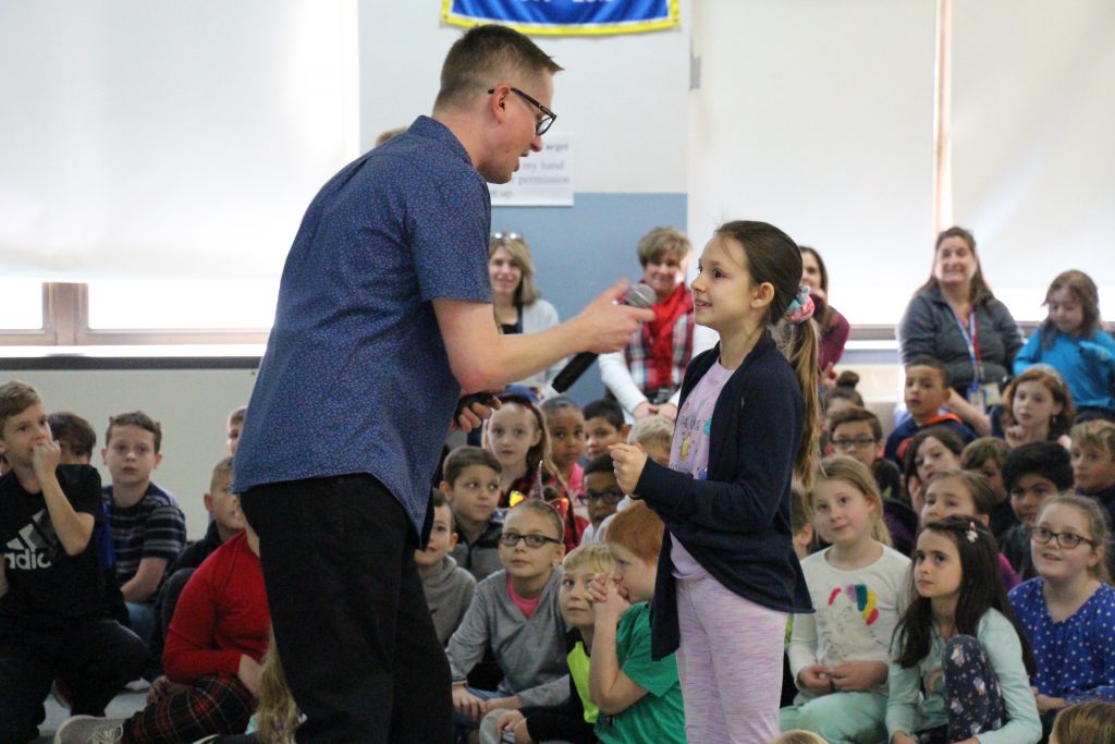 A man in a blue shirt holds a microphone for a young girl who is telling him about books she likes. Many children are in the background.