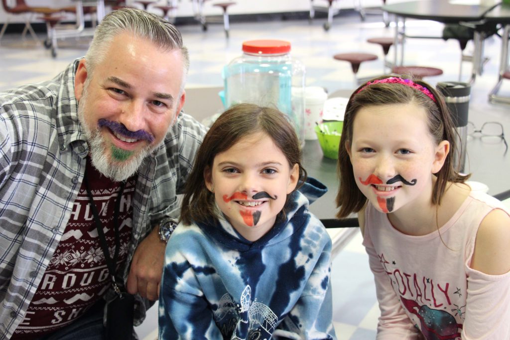 A man with a beard and mustache wearing a flannet shirt sits with two students who have mustaches drawn on their faces.