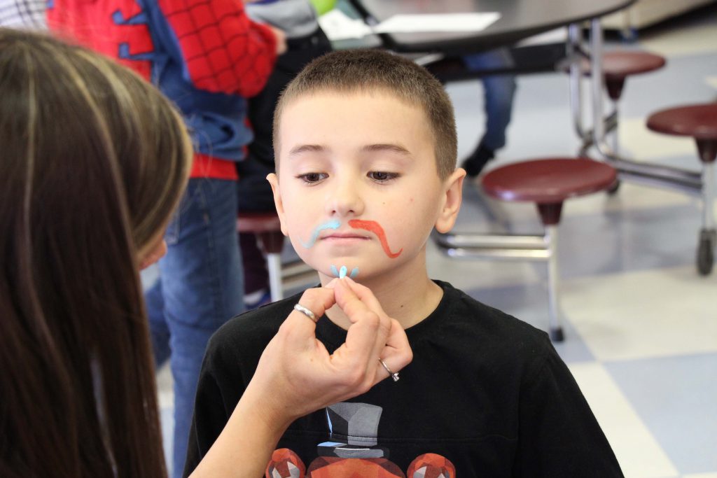An elementary school boy wearing a black shirt has a mustache painted on his face.