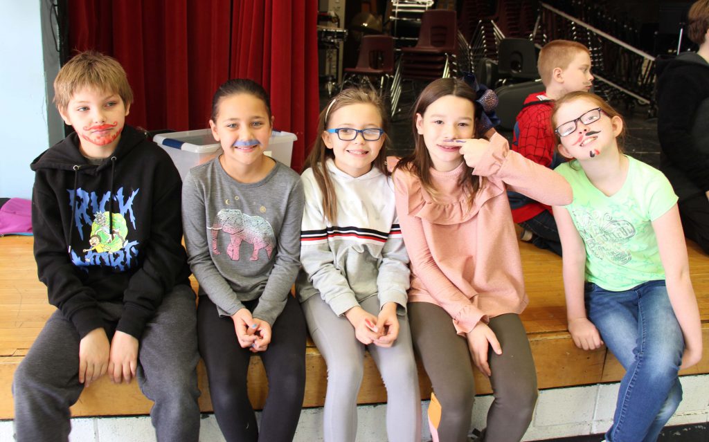 Five elementary students sit on the stage, all with painted mustaches.