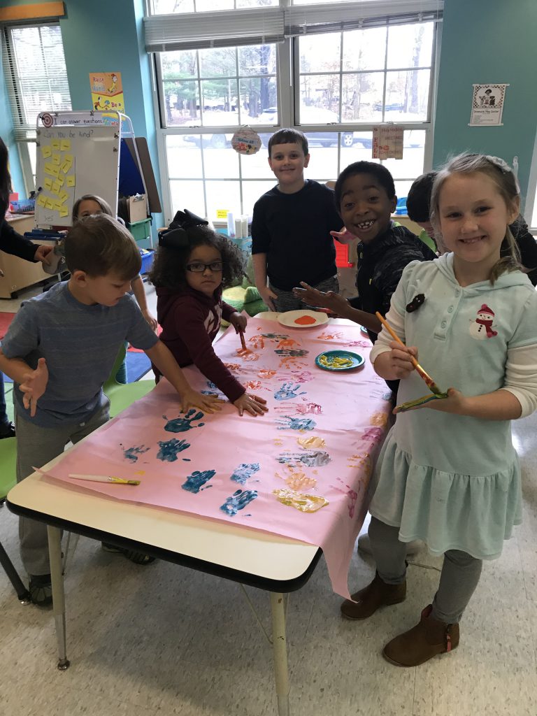 A group of six elementary school children stand around a table that has a large piece of paper on it. The are putting their handprints in paint on it.