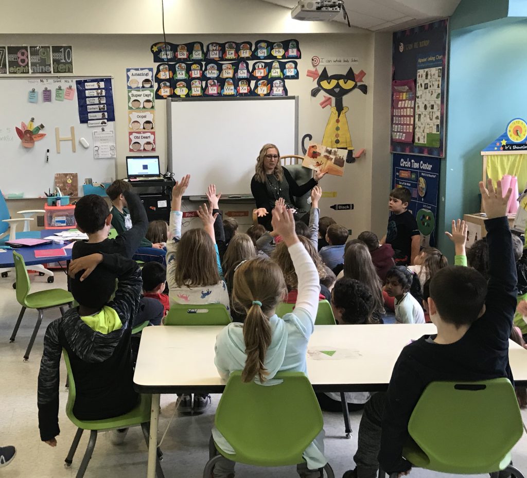 A teacher sits at the front of a class reading from a book, holding it up so the students can see it. The students are sitting on the run as well as in chairs, many with their hands up.