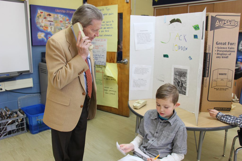 A man in a tan suit jacket holds a phone to his ear as a third-grade boy recites information to him.