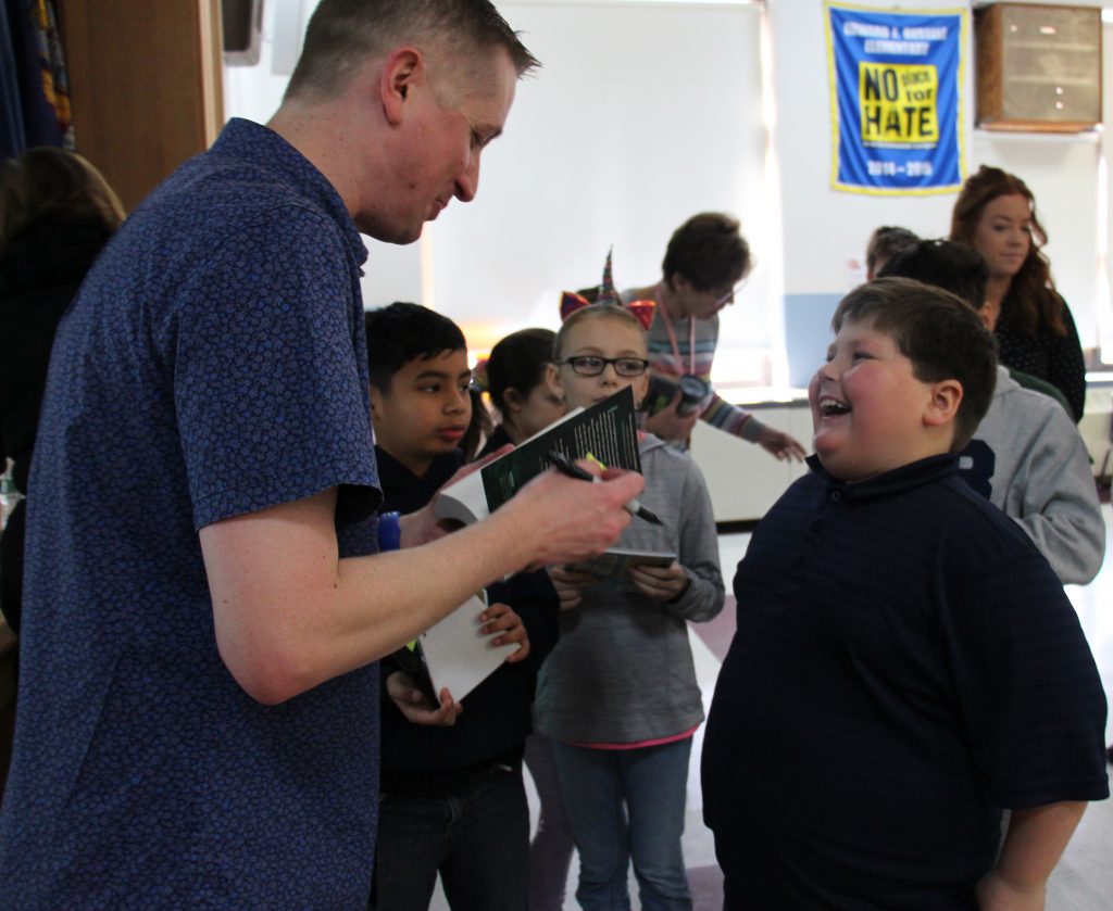 A boy smiles broadly as the man in the blue shirt signs a copy of a book