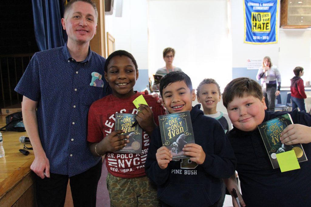 A man in a blue shirt stands with three elementary school boys, all holding books.