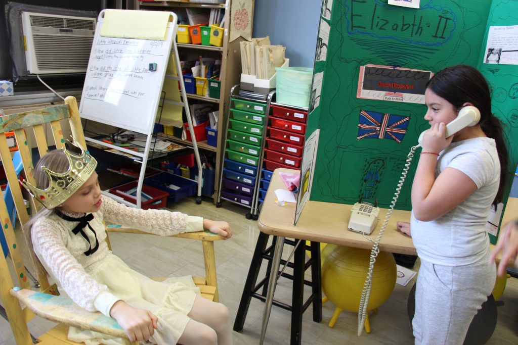 A girl dressed as Queen Elizabeth II talks to a woman holding a phone.