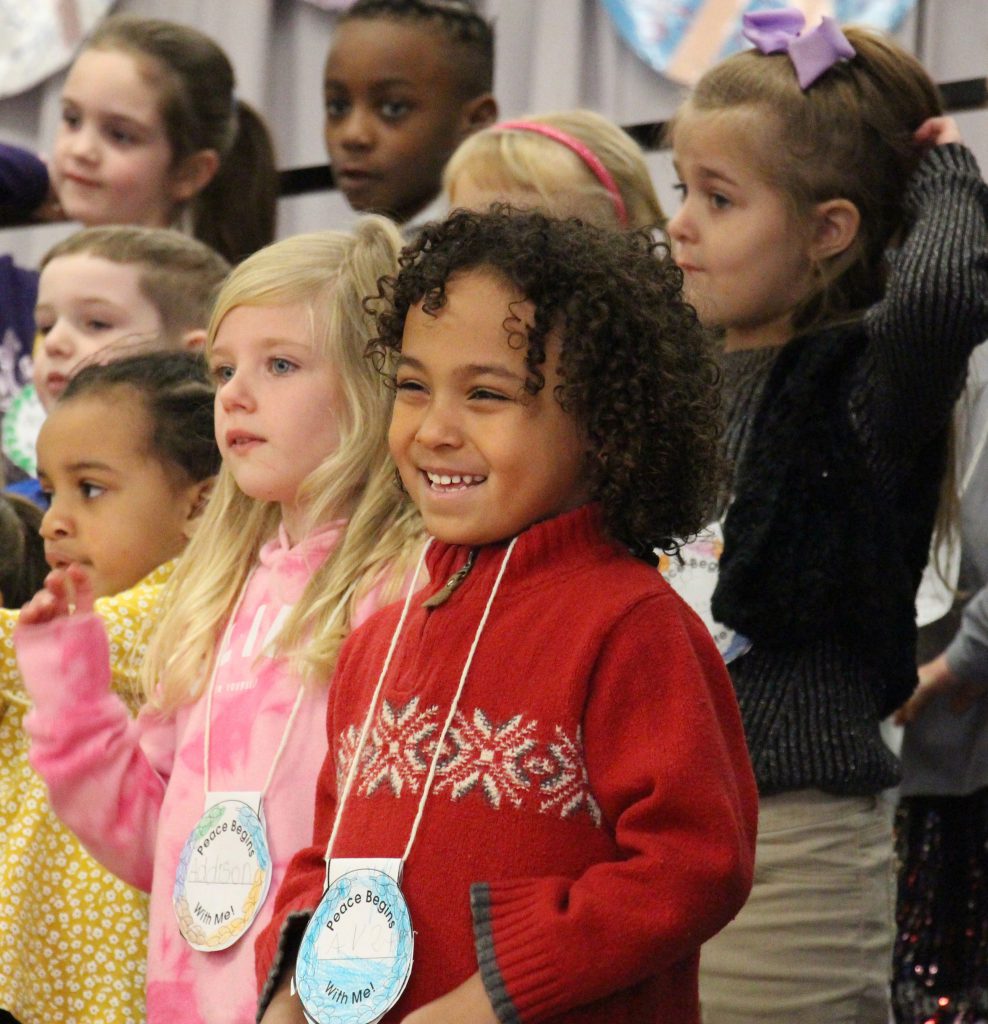 A kindergarten student with shoulder-length curly hair smiles broadly while singing in the concert. He is wearing a bright red sweater.