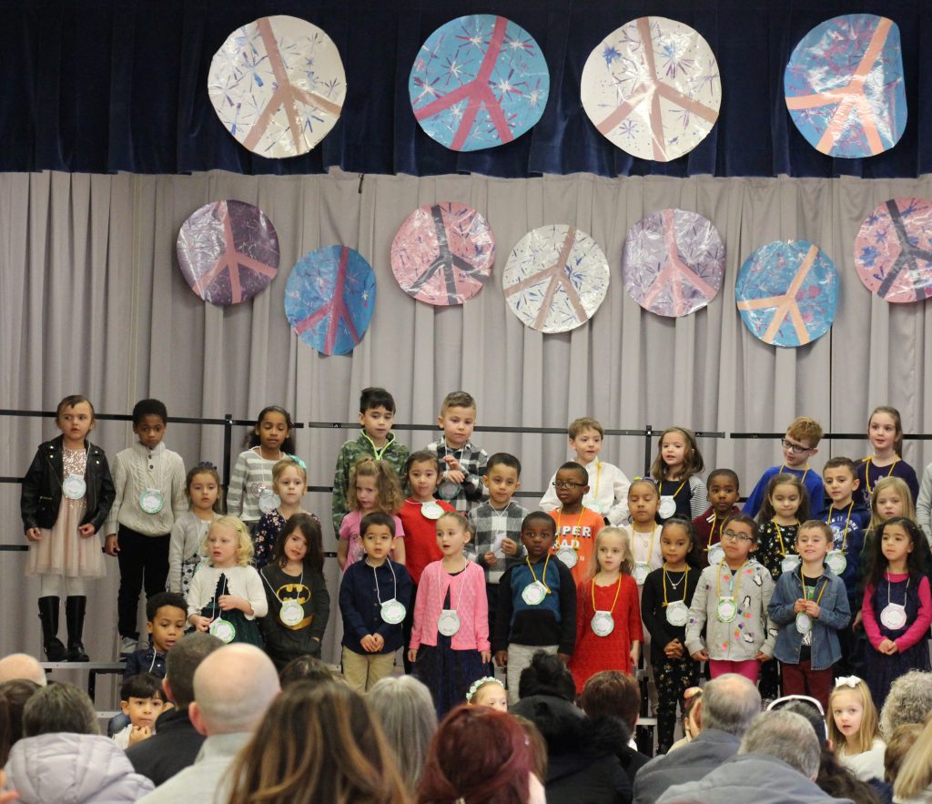 The stage is decorated with large peace signs. About 35 kindergarten students are on the stage singing.