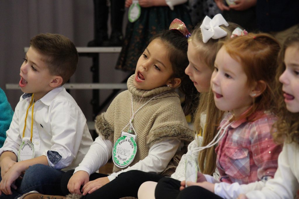 Four kindergarten students - three girls and one boy - sit on the stage singing.