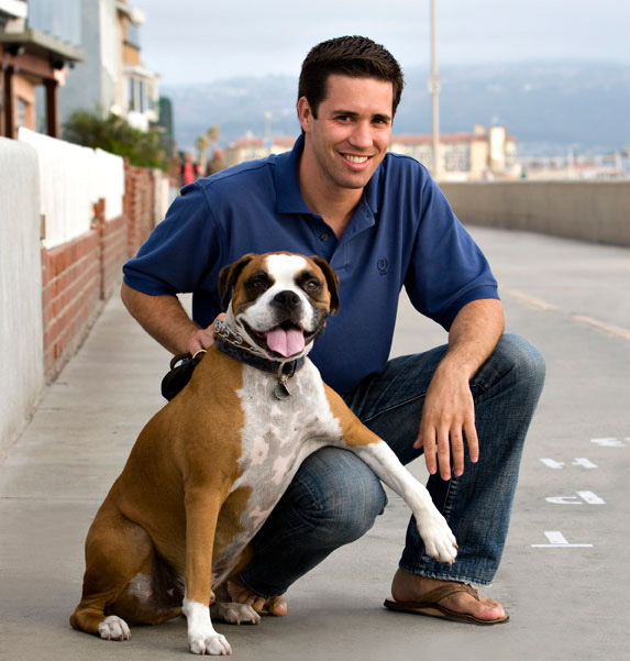 A man wearing a blue, short-sleeve shirt crouches down next to a brown and white dog