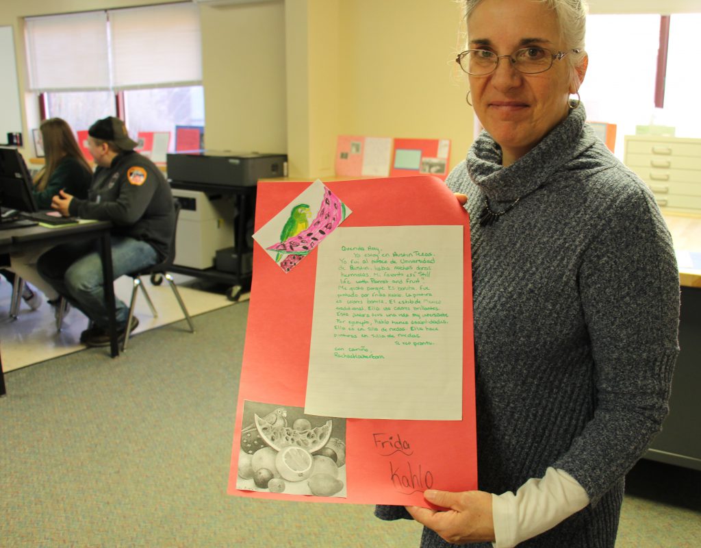 A woman with glasses wearing a gray shirt holds a poster with art on it