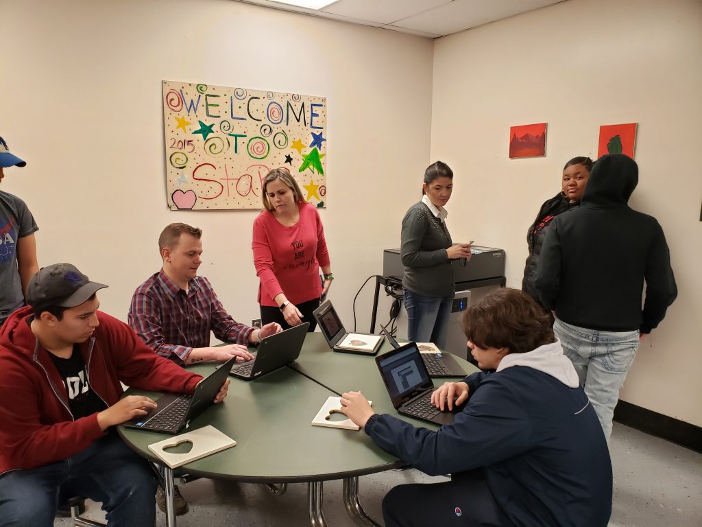 Students sit around a table with their frames, with a teacher leaning in to help them.