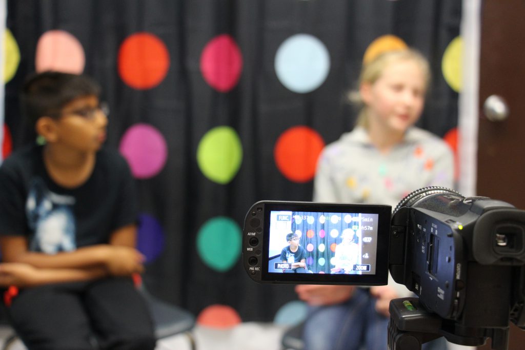 Two students in front of a multi-colored polka dot background. They are seen through the video screen of a camera.