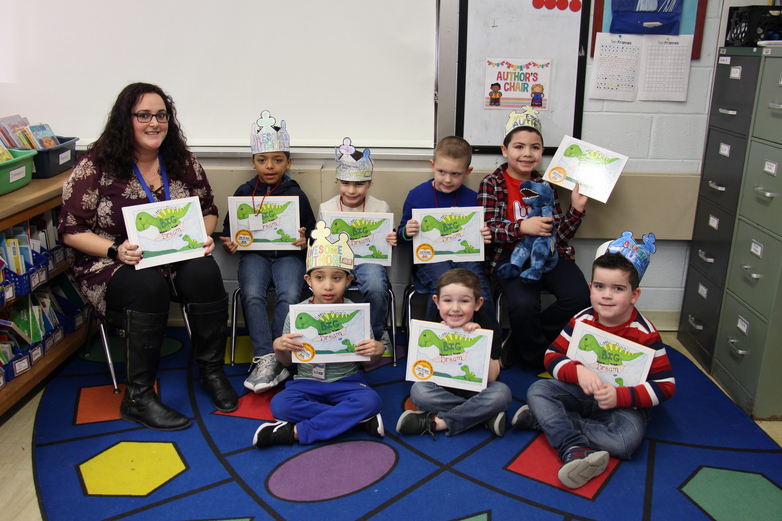 A teacher sits with seven of her young elementary age students. They are all holding up a book they created as a class.