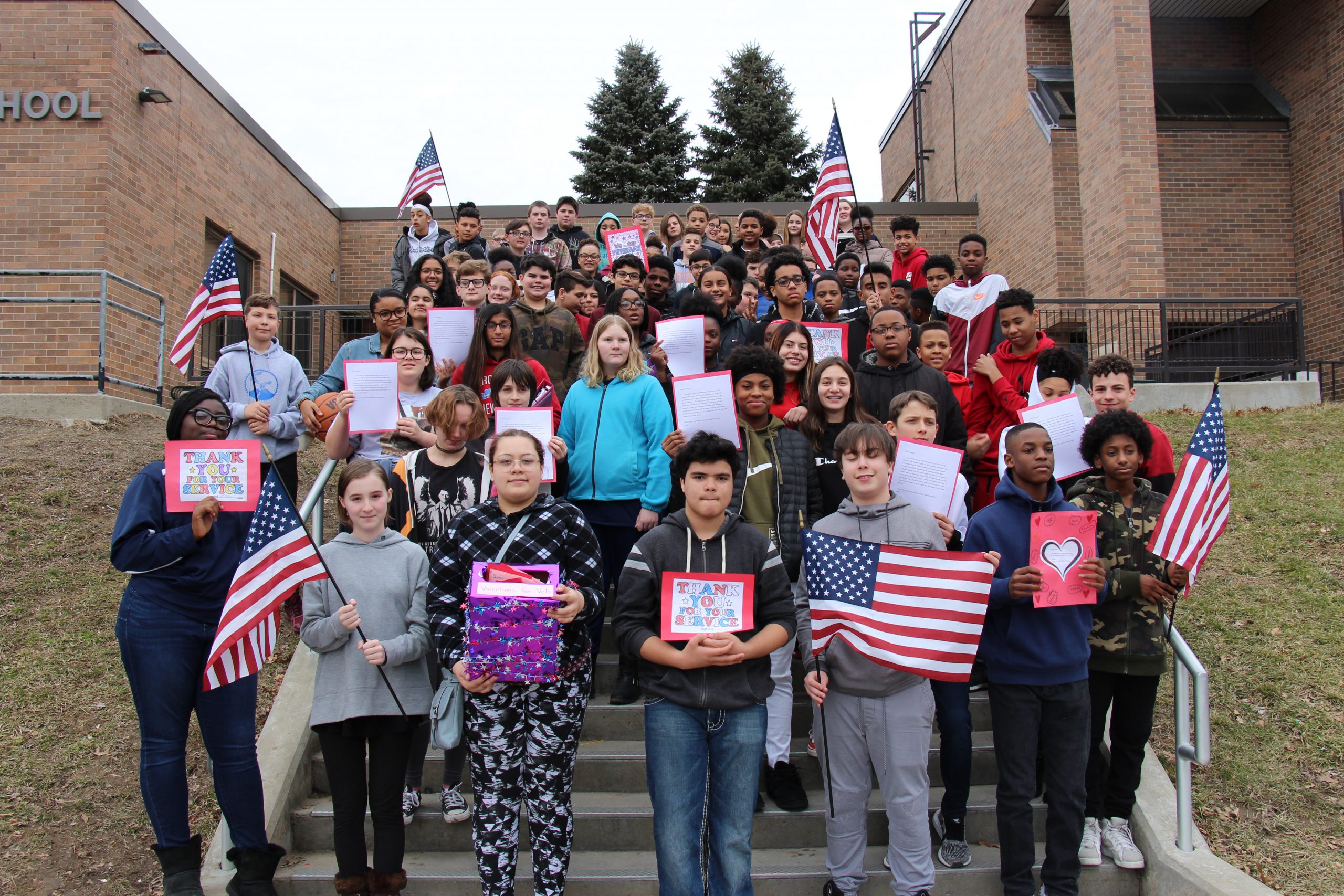 A large group of eighth grade students stand on the steps leading to the school. Many are holding Valentines and American flags.