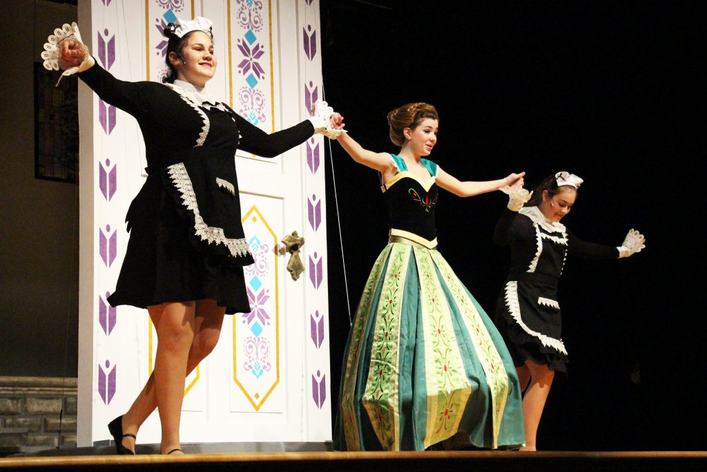 Three young women dance holding hands wide. One is in a ball gown of green, ,gold and black. The others are in maids dresses.