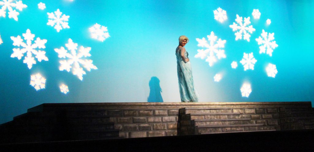 Against a blue backdrop with large snowflakes reflected on it, a young woman dressed in a blue dress stands on a wall.