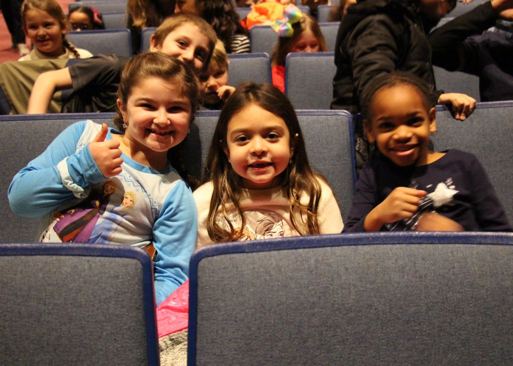 Three elementary age girls smile and give thumbs up sitting in their seats.