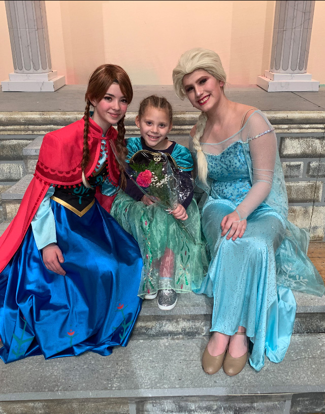 The two princess characters dressed in their gowns sit on the steps with a young girl, also dressed in a princess gown.