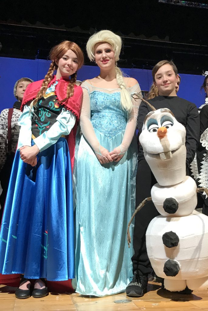 Two middle school girls and a middle school boy stand smiling in their costumes. The two girls are in gowns and the boy is holding a large snowman puppet.