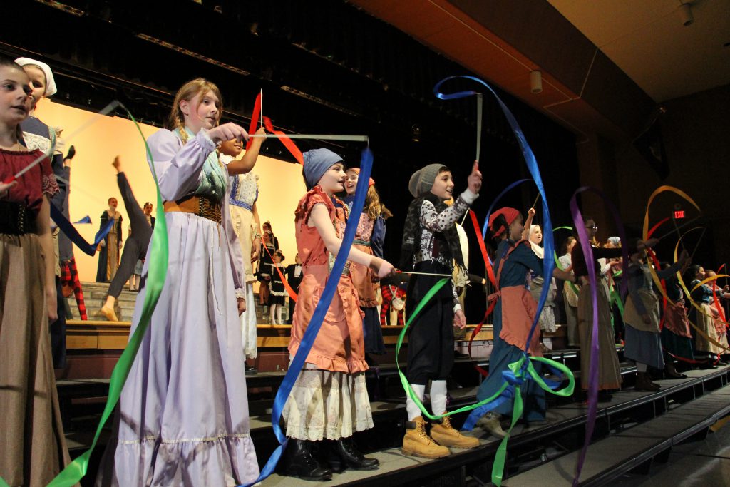 Middle school girls and boys dressed in costumes twirl brightly colored ribbons on sticks.