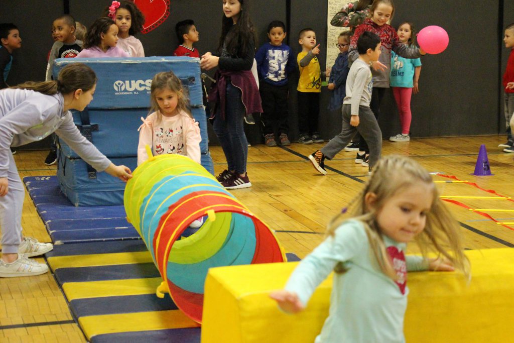 Two little girls on an obstacle course, one jumping over a yellow block and another looking into a multi-colored tunnel.