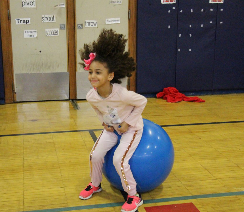 A girl in pink pants and shirt with a red bow in her hair bounces on a blue hippity-hoppity ball.