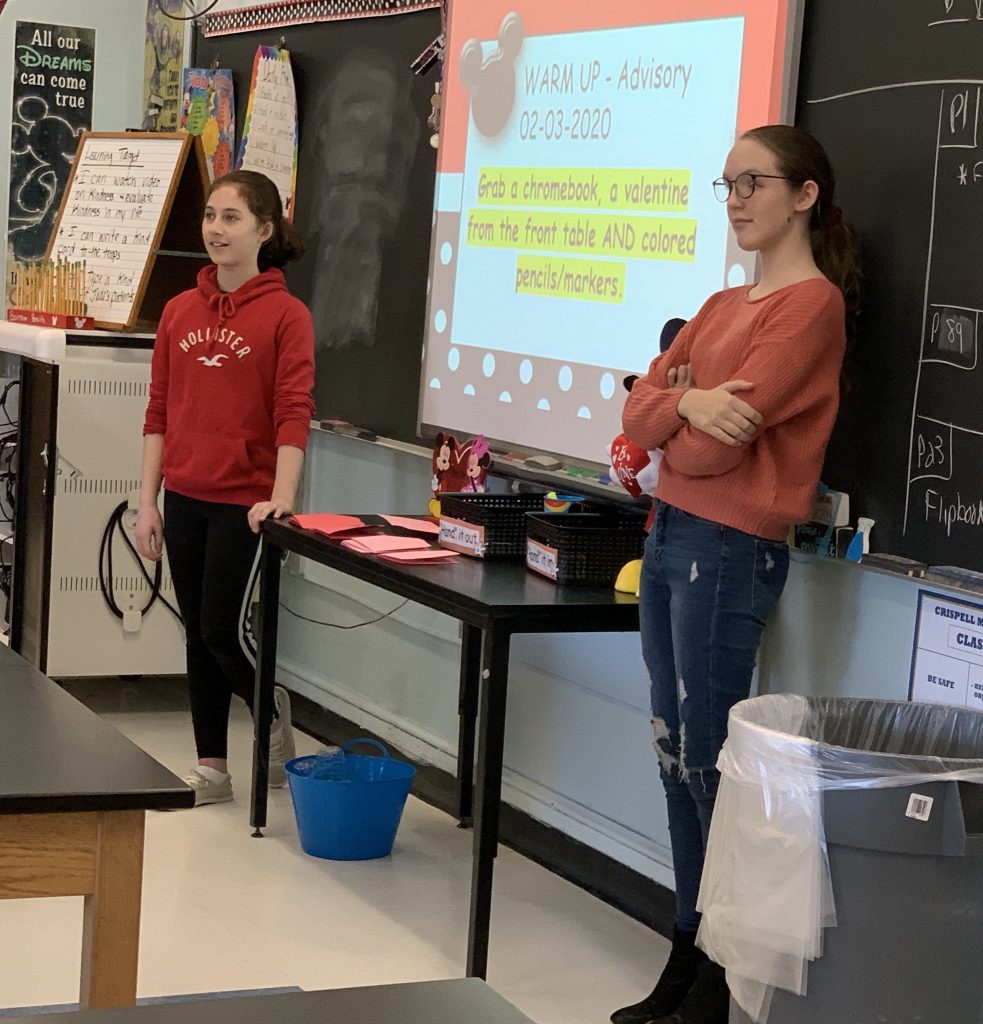 Two middle school students stand on either side of a screen with instructions on sending Valentines.