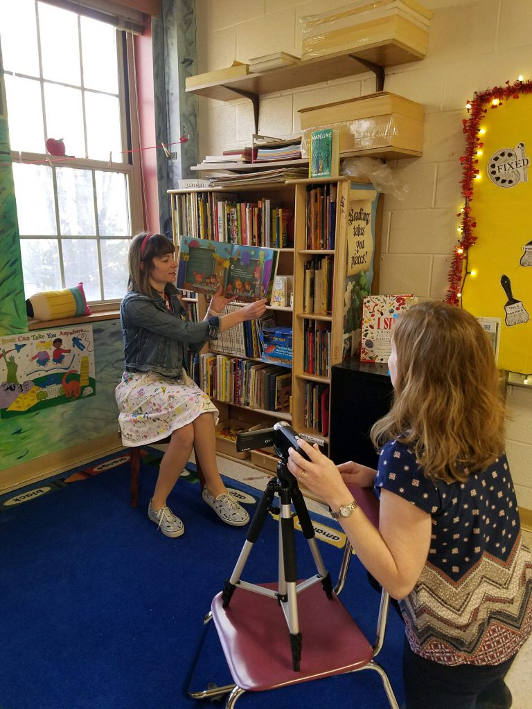 A woman sits on a chair holding a book up and reading it as another woman videos her. It is in a classroom.