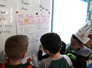 Three elementary age boys write names of the books they wrote on a white board.