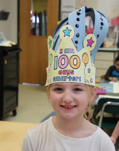 A little girl smiles and wears a paper crown with the number 100 on it 