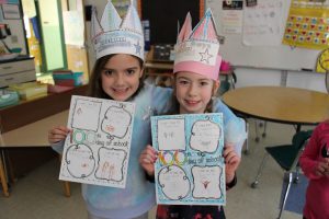 Two elementary school girls wear paper hats and hold up a list of things having to do with the number 100