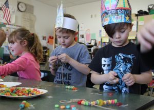 Kindergarten students wearing paper crowns string colorful cereal to create necklaces.