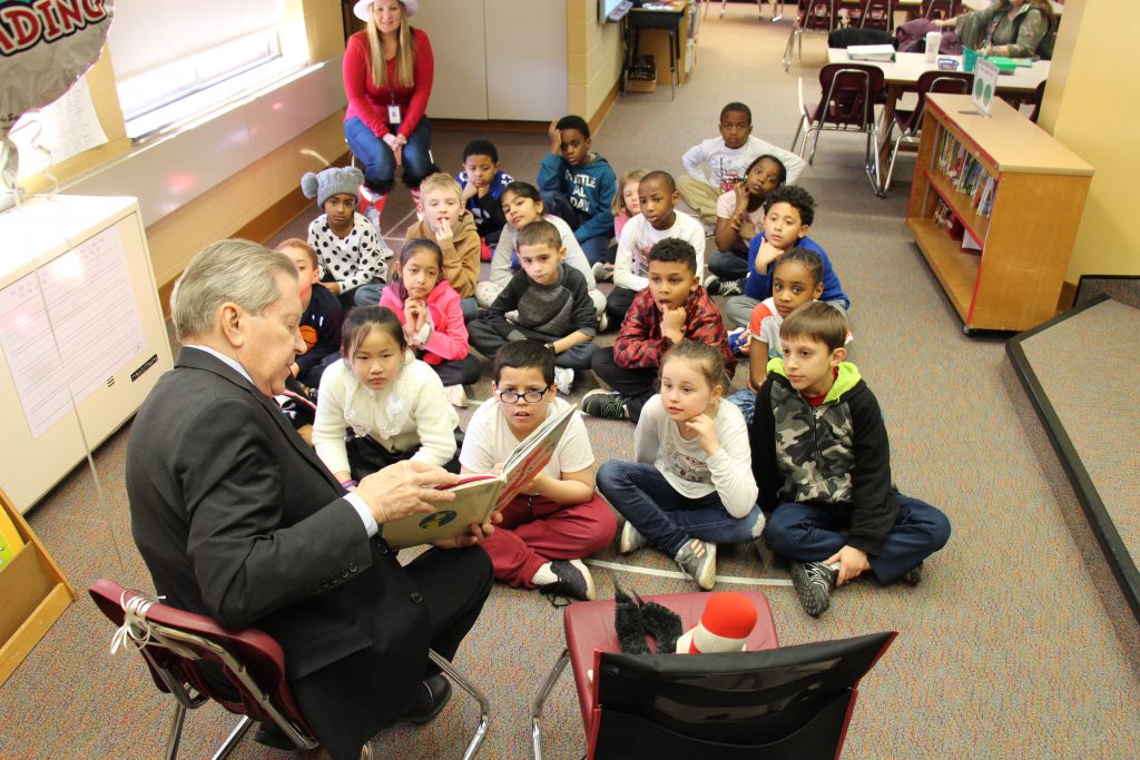 A class of second grade students sit on a rug in the library and listen as a man reads Fox in Sox to them.