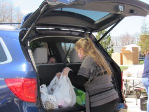 A woman with long blonde hair puts white plastic bags of food in the trunk of a blue car.
