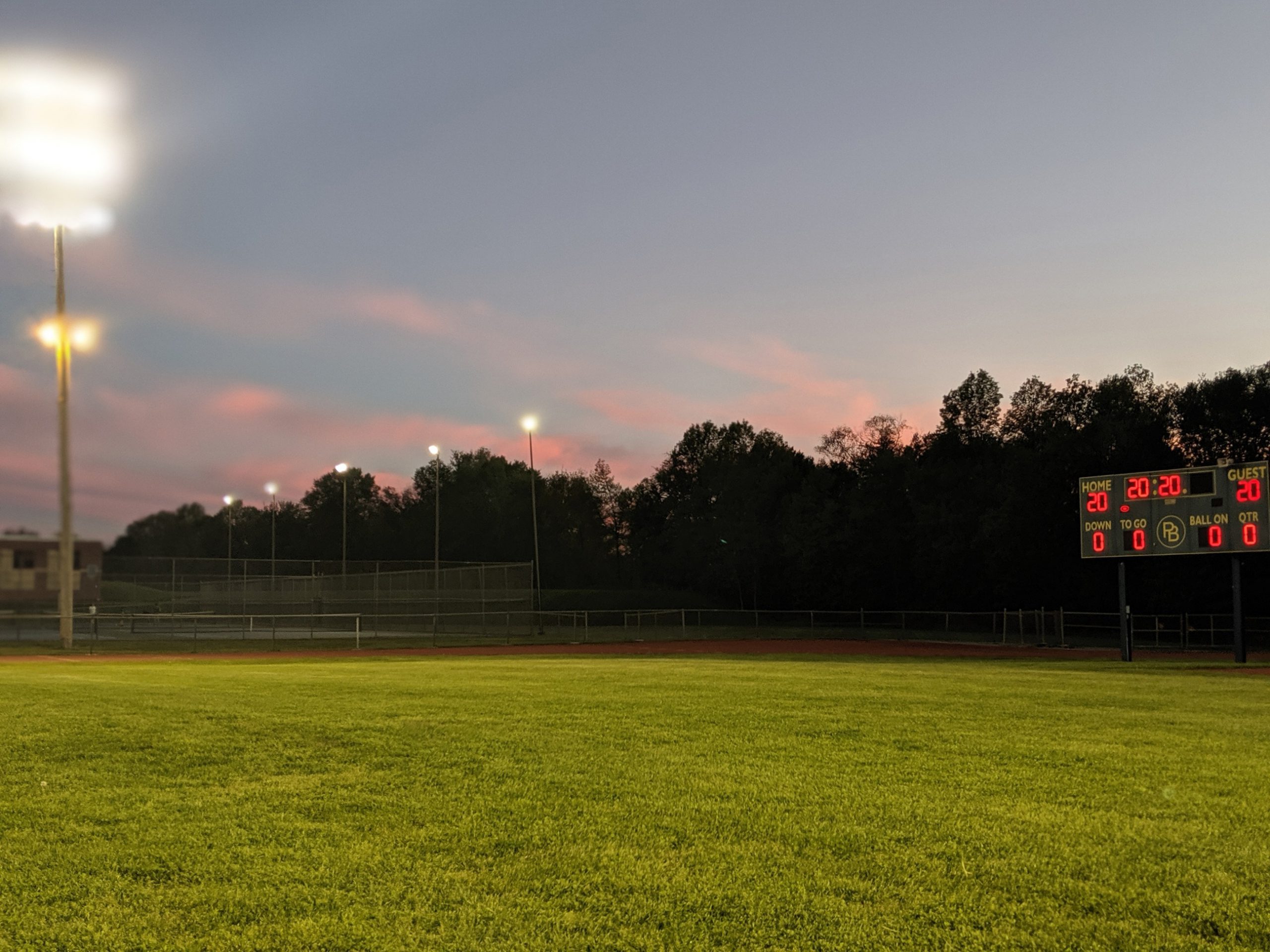 The sky at dusk, football field grass with the scoreboard  lit saying 20 20 and the tower lights are on